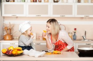 Little cute girl with her mother eating orange while cooking. Kitchen interior. Concept for young kitchen hands